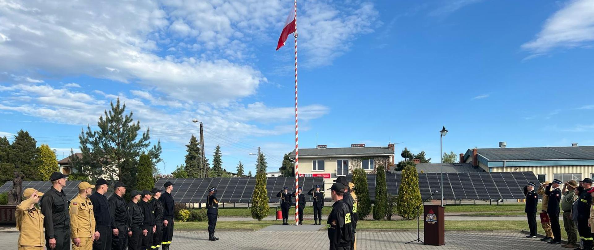 Na pierwszym planie, po prawej i lewej stronie w dwóch rzędach stoją strażacy w umundurowaniu koszarowym. Na wprost flaga państwowa wciągnięta na maszt, za nią poczet flagowy. Osoby funkcyjne oddają honory.