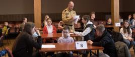 Na zdjęciu dzieci siedzą przy stolikach, funkcjonariusz straży pożarnej rozdaje kartki z testami.