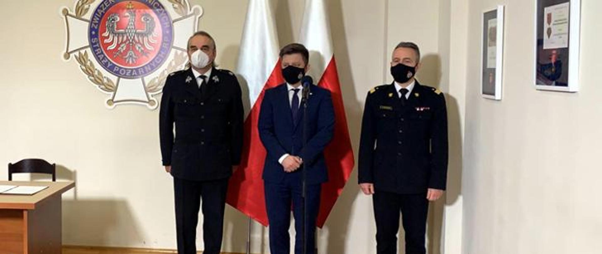 Zdjęcie przedstawia 3 osoby w pozycji stojącej obok siebie. Zdjęcie wykonane wewnątrz budynku na tle białej ściany. Za osobami znajduje się logo Związku Ochotniczych Straży Pożarnych RP oraz flaga Polski.