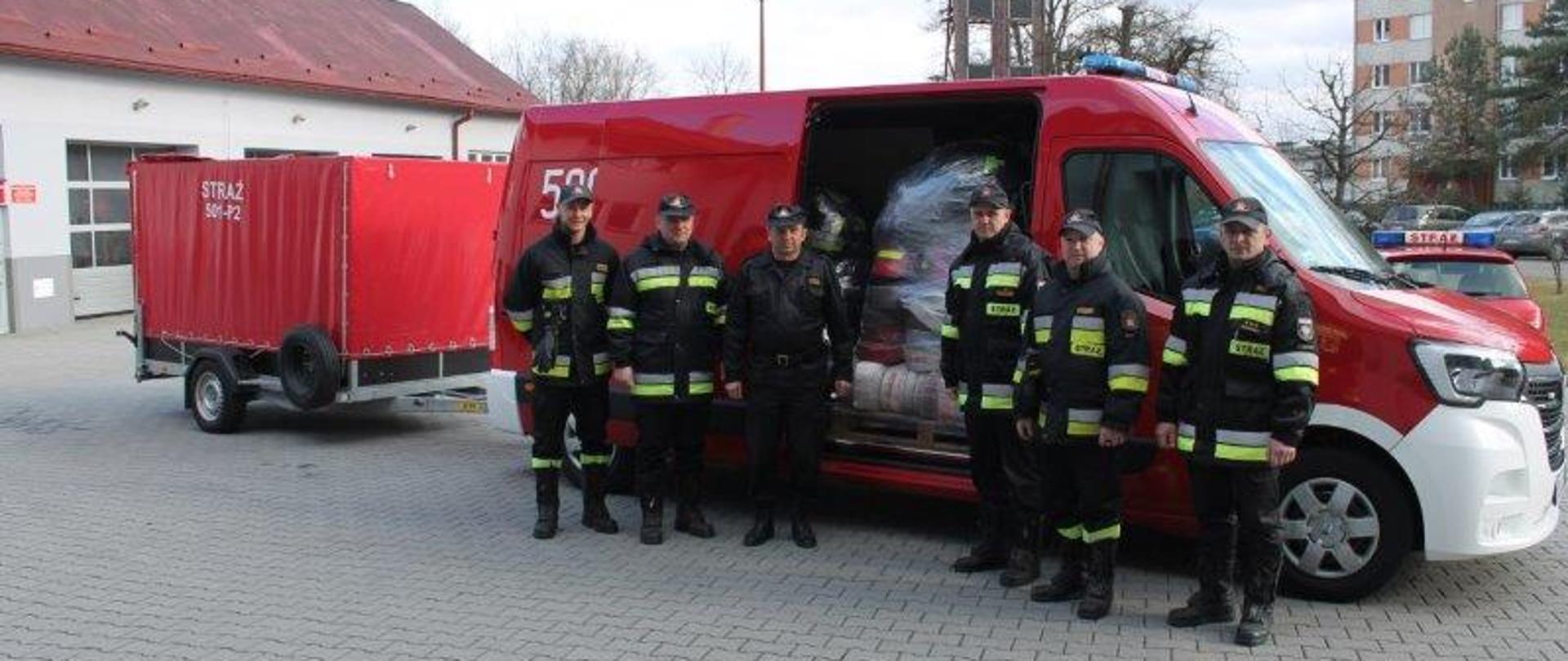 Na fotografii plac komendy powiatowej Państwowej Straży Pożarnej w Mielcu. Na placu widać funkcjonariuszy komendy powiatowej PSP w Mielcu na tle samochodu z przyczepką załadowanych sprzętem zebranym w powiecie mieleckim dla Ukraińskich strażaków. 