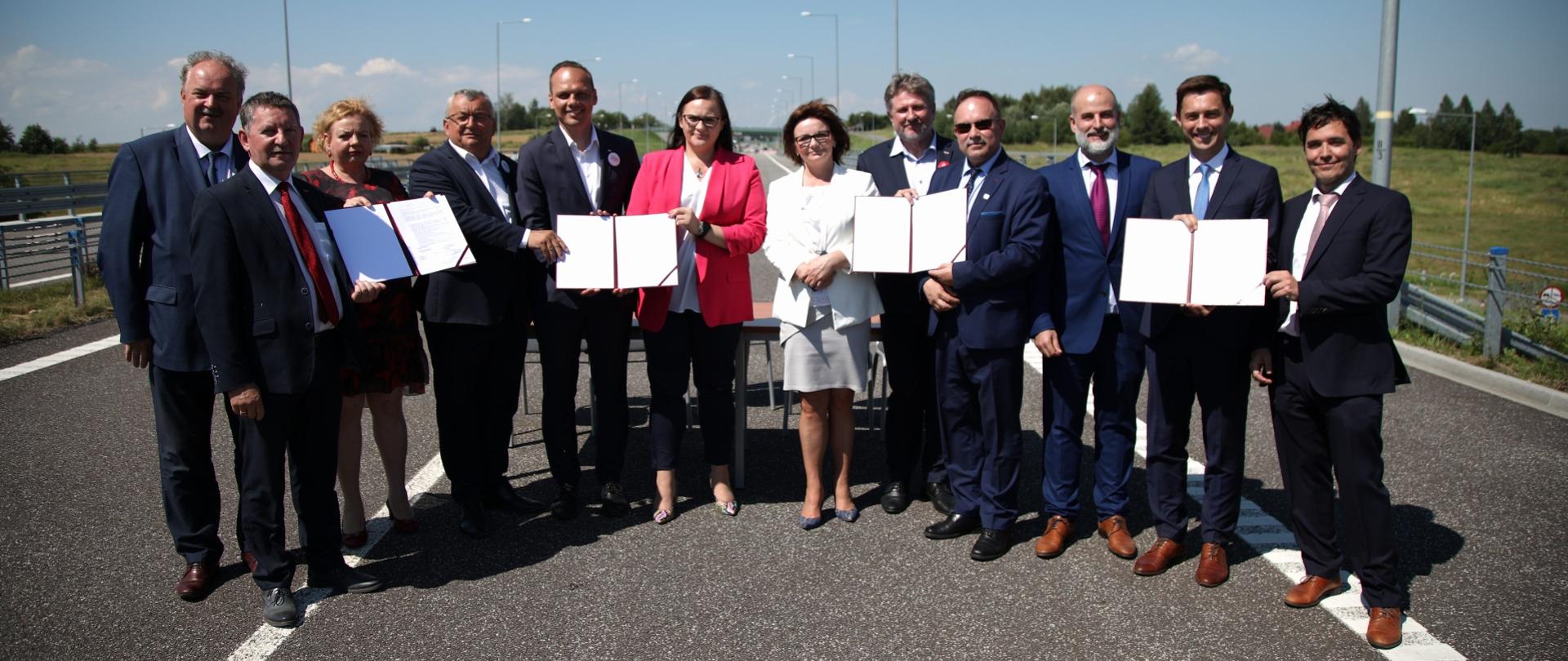 Na drodze asfaltowej stoi grupa ludzi z dokumentami w rękach. Pośrodku minister Małgorzata Jarosińska-Jedynak.