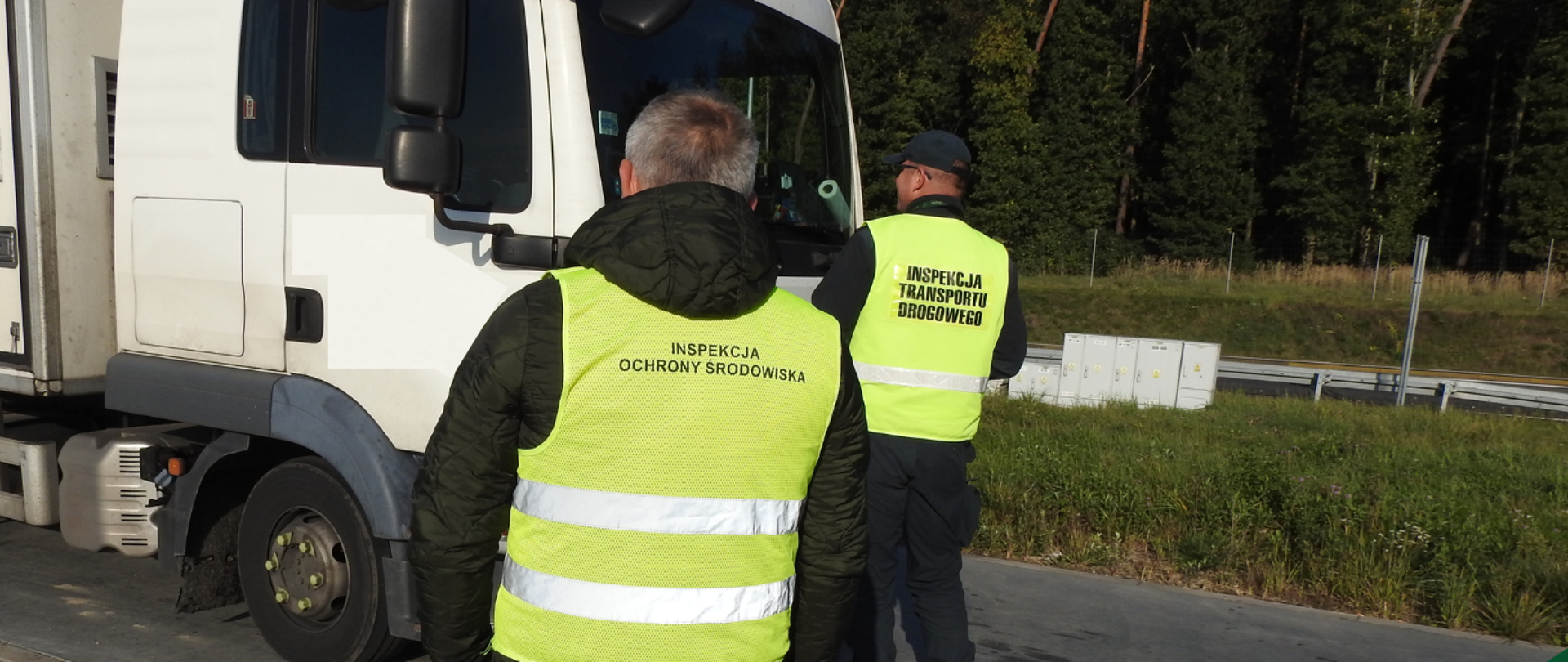 Dwóch mężczyzn w odblaskowych kamizelkach. Są oni obróceni tyłem do zdjęcia, mężczyzna na pierwszym planie ma na kamizelce napis Inspekcja Ochrony Środowiska, a mężczyzna na drugim planie ma napis Inspekcja Transportu Drogowego. Stoją przy samochodzie ciężarowym na poboczu przy lesie.