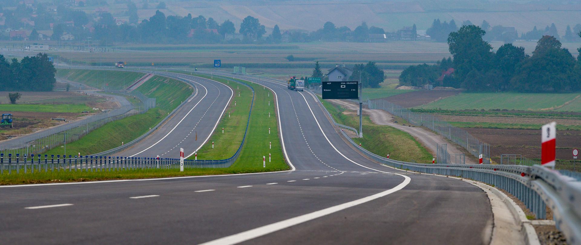 Nowo wybudowany odcinek dwujezdniowej drogi ekspresowej z pasem zieleni pośrodku, barierami ochronnymi i oznakowaniem. Droga biegnie przez otwarty, wiejski krajobraz z polami i nielicznymi zabudowaniami w tle, przy lekko zamglonym niebie.