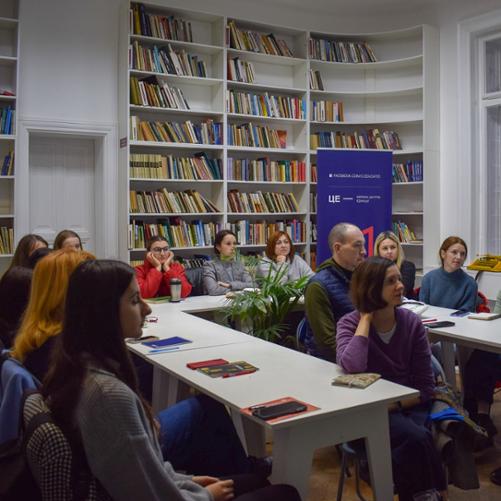 W pomieszczeniu z regałami wypełnionymi książkami grupa osób siedzi przy stołach ustawionych w podkowę i patrzy się na prowadzącego zajęcia