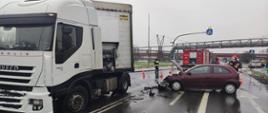samochód osobowy z rozwlonym przodem w poprzek drogi przy samochodzie ciężarowym z naczepą. W tle samochód straży oraz strażacy