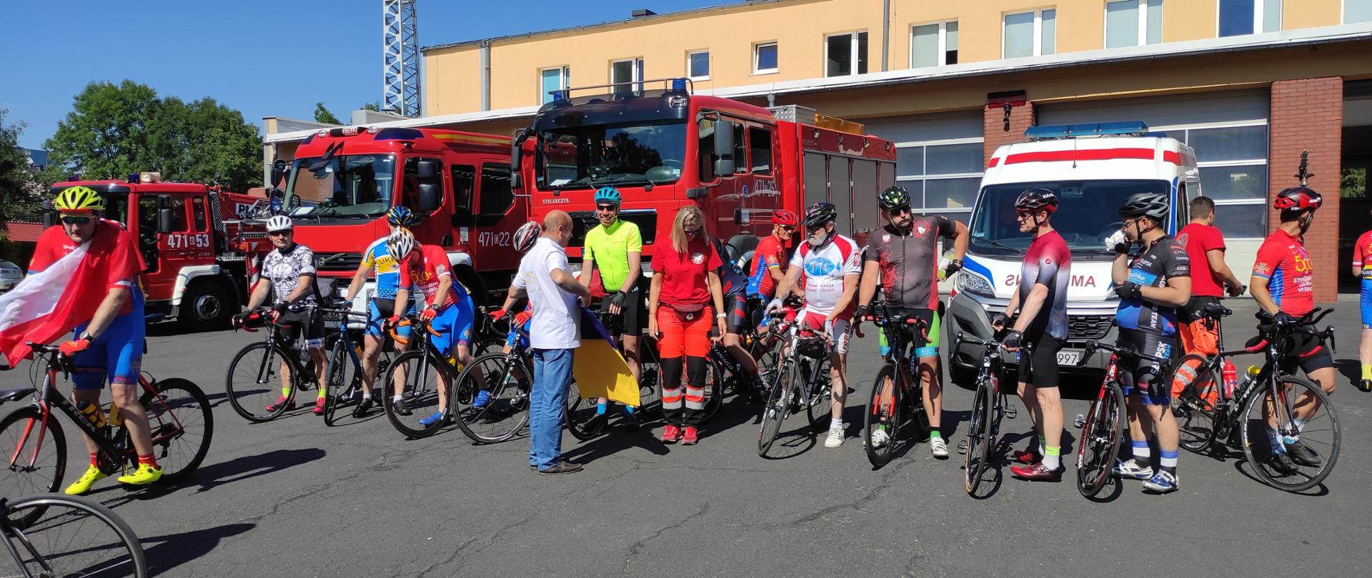 Przed garażami strażackimi stoją samochody pożarnicze oraz karetka pogotowia, a przed samochodami kolarze z rowerami.