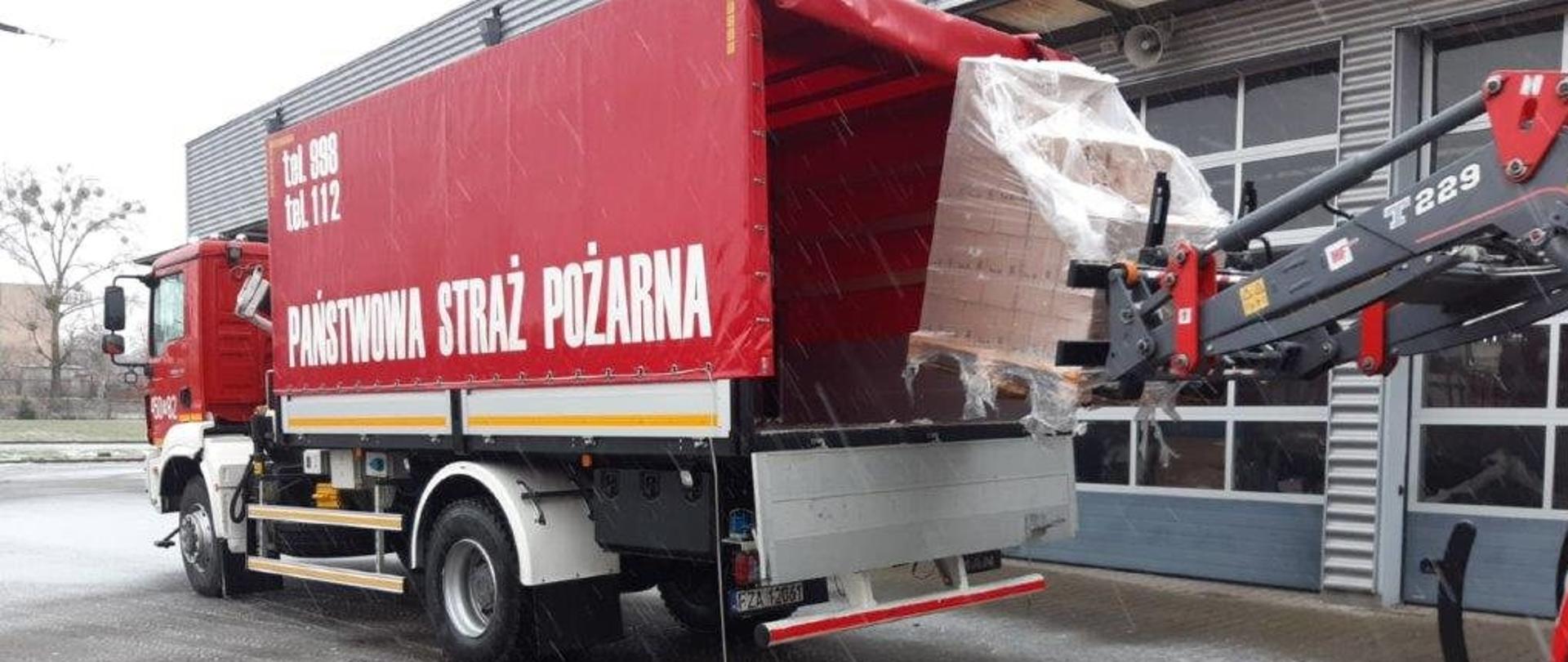 Zdjęcie przedstawia samochód ciężarowy PSP z otwartą tylną klapą. Do samochodu podjeżdża ciągnik z podnośnikiem widłowym, z paletą ze środkami do dezynfekcji.