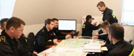 Funkcjonariusze Państwowej Straży Pożarnej siedzą za stołem na którym rozłożone są mapy niedaleko stoi monitor oraz tablica na której wyznaczone są siły i środki. 