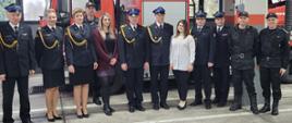 Strażacy i pracownicy cywilni wraz z kończącymi służbę strażakami stoją na tle drabiny pożarniczej. 