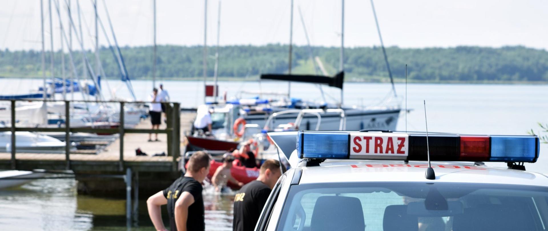 Zdjęcie wykonane na zewnątrz na pierwszym planie belka samochodu strażackiego z napisem straż z tyłu jezioro oraz keja, przy której zacumowane są żaglówki.