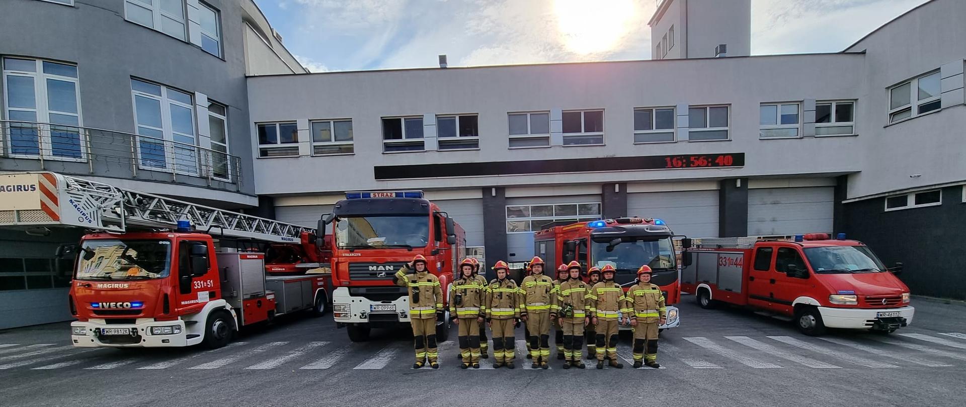 Zdjęcie wykonane przed jednostką ratowniczo – gaśniczą numer 1 w Radomiu. Na zdjęciu widać strażaków ustawionych w szeregu "na baczność" przed bramami garażowymi. Strażacy ubrani w ubrania specjalne w kolorze żółtym. Na głowach mają czerwone hełmy. Za strażakami znajdują się pojazdy ratowniczo - gaśnicze. Samochody w kolorze czerwonym z białymi oraz srebrnymi elementami zabudowy. Pojazdy mają zapalone niebieskie światła alarmowe oraz włączone sygnały dźwiękowe. Jeden ze strażaków - dowódca oddaje honory w celu upamiętnienia poległych w Powstaniu Warszawskim.
