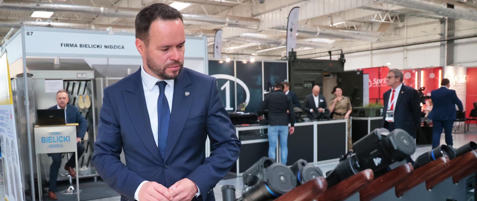 Wojewoda Lubelski Krzysztof Komorski, w granatowym garniturze, z uwagą ogląda broń snajperską i inny sprzęt optyczny prezentowany na stoisku podczas VIII edycji Targów „GRANICE” w Lublinie. W tle widoczne są inne stoiska wystawców, z widocznym napisem „FIRMA BIELICKI NIDZICA” oraz ogólna przestrzeń targowa z licznymi uczestnikami, co podkreśla różnorodność oferty firm zbrojeniowych i zaawansowanych technologii dla ochrony granic. Zainteresowanie wojewody ekspozycją świadczy o ważności modernizacji służb i wykorzystania nowoczesnych rozwiązań.