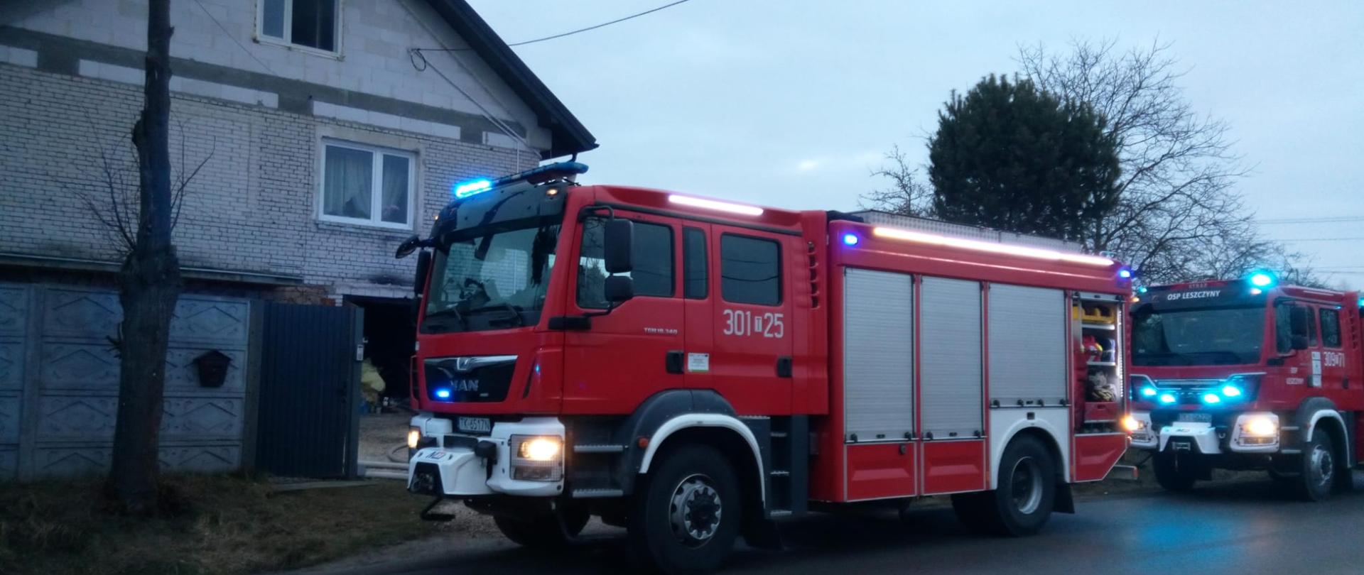 Zdjęcie przedstawia ulicę na której stoją dwa samochody pożarnicze. Głębiej, po lewej stronie, za betonowym ogrodzeniem widać dom jednorodzinny w którym doszło do pożaru. Brama garażowa otwarta a do wnętrza wprowadzone są węże gaśnicze. Widać okopcone nadproże nad bramą garażu.