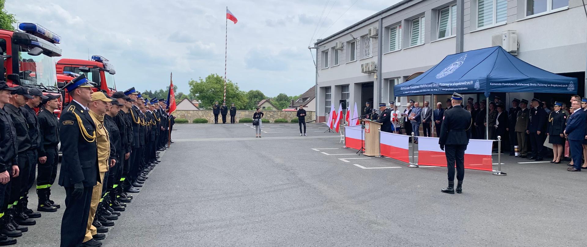 Zdjęcie przedstawiające uroczystość Dnia Strażaka w KP PSP Krapkowice. Po lewej na tle ustawionych pojazdów pożarniczych stoi pododdział strażaków PSP i OSP. W środku w oddaleniu widać maszt flagowy z poniesioną flaga RP i stojący u jego stóp poczet flagowy. Po prawej na tle zabudowań komendy ustawiona trybuna honorowa, z której przemawia Komendant Powiatowy PSP. Pod niebieskim namiotem za komendantem stoją zaproszeni goście. Bliżej przed trybuną, tyłem do patrzącego ustawiony dowódca uroczystości. 