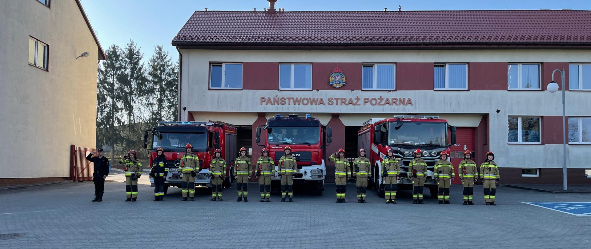 Na miejscu widoczni strażacy na tle samochodów pożarniczych oraz budynku jednostki