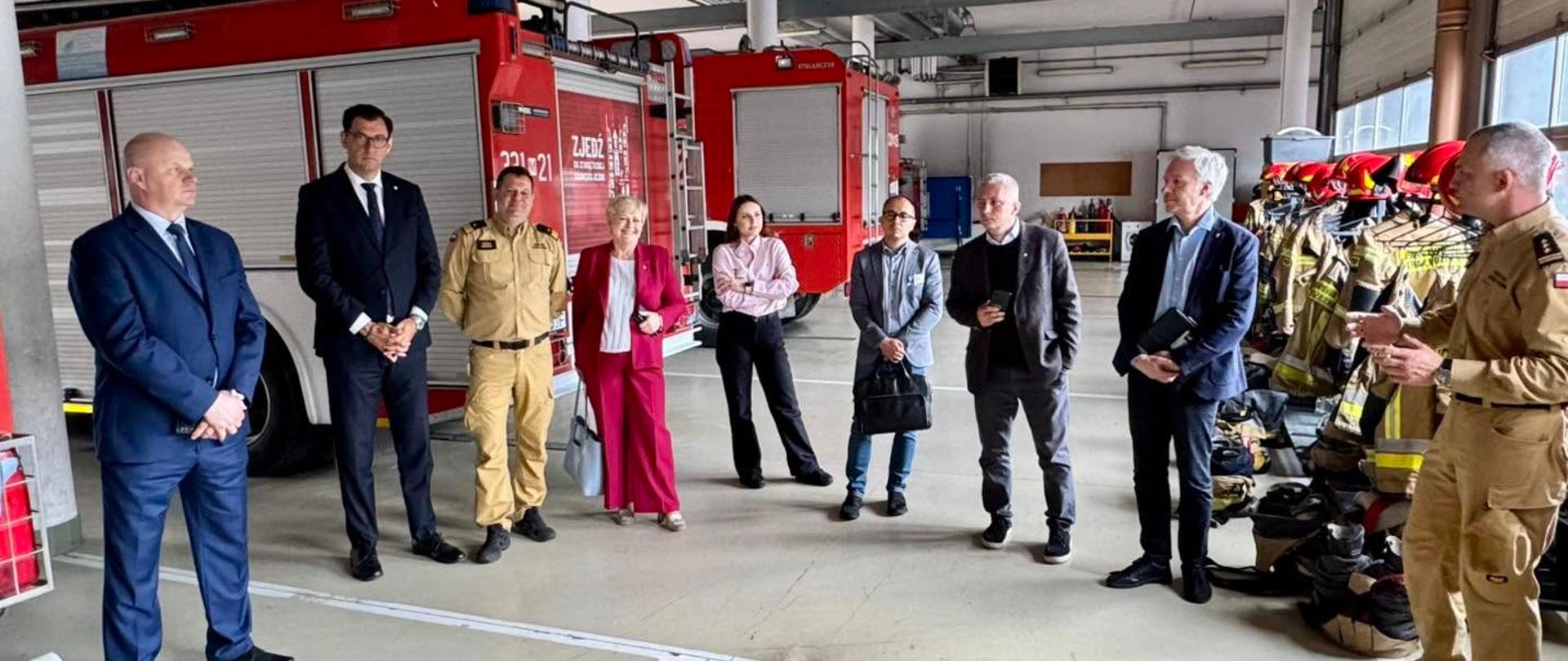 Na zdjęciu przedstawiono spotkanie delegacji sejmowej Komisji Administracji i Spraw Wewnętrznych z przedstawicielami Państwowej Straży Pożarnej wewnątrz garażu Jednostki Ratowniczo-Gaśniczej nr 1 w Radomiu. W centralnej części kadru stoi grupa kilkunastu osób, wśród których znajdują się posłowie, przedstawiciele administracji publicznej oraz funkcjonariusze PSP. Strażacy ubrani są w mundury służbowe koloru piaskowego, natomiast pozostali uczestnicy mają na sobie stroje biznesowe i garnitury. Po prawej stronie widoczny jest strażak prezentujący informacje uczestnikom spotkania. W tle znajdują się czerwone samochody pożarnicze, stanowiska garażowe oraz elementy wyposażenia jednostki ratowniczo-gaśniczej. Zdjęcie wykonano wewnątrz budynku, w dobrze oświetlonym garażu.