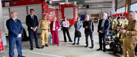 Na zdjęciu przedstawiono spotkanie delegacji sejmowej Komisji Administracji i Spraw Wewnętrznych z przedstawicielami Państwowej Straży Pożarnej wewnątrz garażu Jednostki Ratowniczo-Gaśniczej nr 1 w Radomiu. W centralnej części kadru stoi grupa kilkunastu osób, wśród których znajdują się posłowie, przedstawiciele administracji publicznej oraz funkcjonariusze PSP. Strażacy ubrani są w mundury służbowe koloru piaskowego, natomiast pozostali uczestnicy mają na sobie stroje biznesowe i garnitury. Po prawej stronie widoczny jest strażak prezentujący informacje uczestnikom spotkania. W tle znajdują się czerwone samochody pożarnicze, stanowiska garażowe oraz elementy wyposażenia jednostki ratowniczo-gaśniczej. Zdjęcie wykonano wewnątrz budynku, w dobrze oświetlonym garażu.