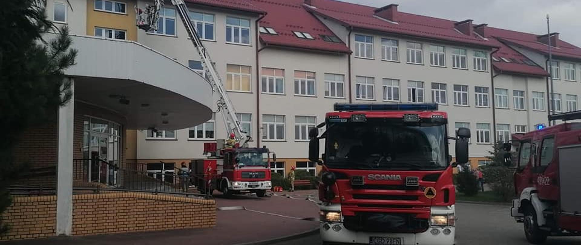 NA ZDJĘCIU WIDAĆ TRZY WOZY STRAŻACKIE, JEDEN Z NICH TO PODNOŚNIK STRAŻACKI, JEST ROZSTAWIONY A JEGO KOSZ ZNAJDUJE SIĘ W POBLIZU BUDYNKU SZKOŁY. W TLE WIDAC BUDYNEK SZKOŁY. 