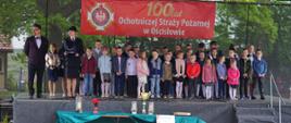 na scenie stoją dzieci. za nimi w tle znajduje się baner z okazji 100 lecia ochotniczej straży pożarnej w ościsłowie