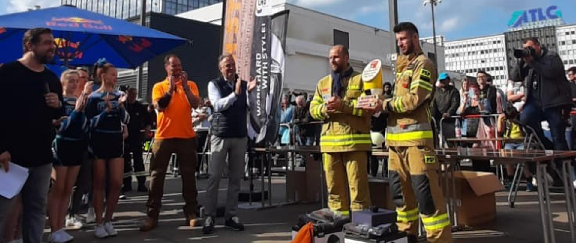 Zdjęcie przedstawia funkcjonariusza komendy miejskiej Państwowej Straży Pożarnej w Radomiu wraz z drugim funkcjonariuszem komendy miejskiej Państwowej Straży Pożarnej w Szczecinie. Zdjęcie wykonane na tle budynku w Berlinie, w którym zostały rozegrane zawody. Strażacy ubrani są w ubrania specjalne strażackie w kolorze piaskowym. Na szyi zawieszone mają pamiątkowe medale. Przed nim ustawiona jest pamiątkowa statuetka oraz nagrody. Dookoła nich ustawieni są inni ludzie, uczestnicy zawodów. Osoby te biją brawo strażakom – zwycięzcom zawodów. 