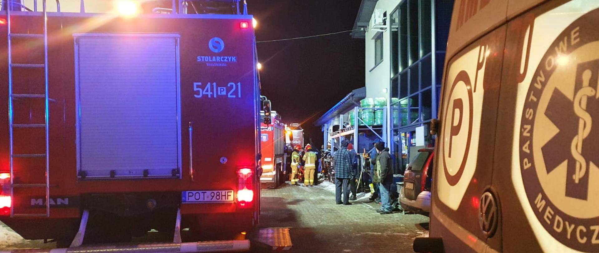 Zdjęcie przedstawia działania ratowniczo - gaśnicze. Na pierwszym planie widać pojazdy ratowniczo- gaśnicze które biorą udział w pożarze w miejscowości Mikstat. Na dalszym planie widzimy ratowników biorących udział w akcji. Zdjęcie wykonane w porze wieczornej przed posesją.