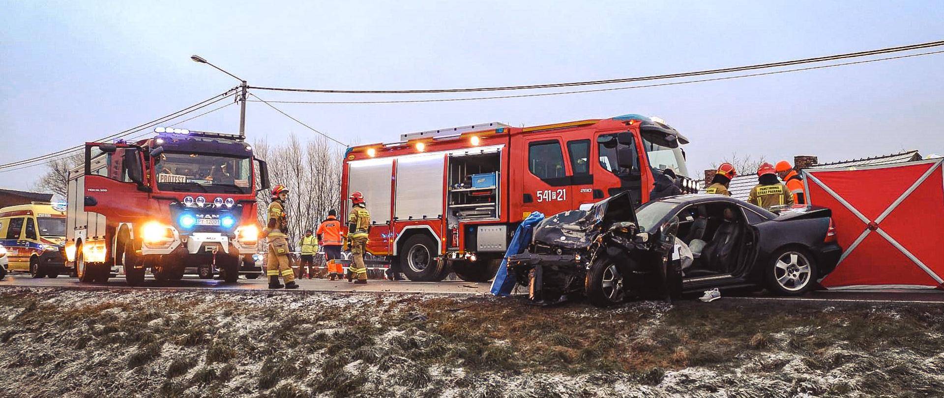 Na pierwszym planie uszkodzony samochód osobowy w wyniku zderzenia z ciężarówką. Po prawej stronie parawan i ratownicy. Na dalszym planie wozy strażackie i ambulans pogotowia ratunkowego.