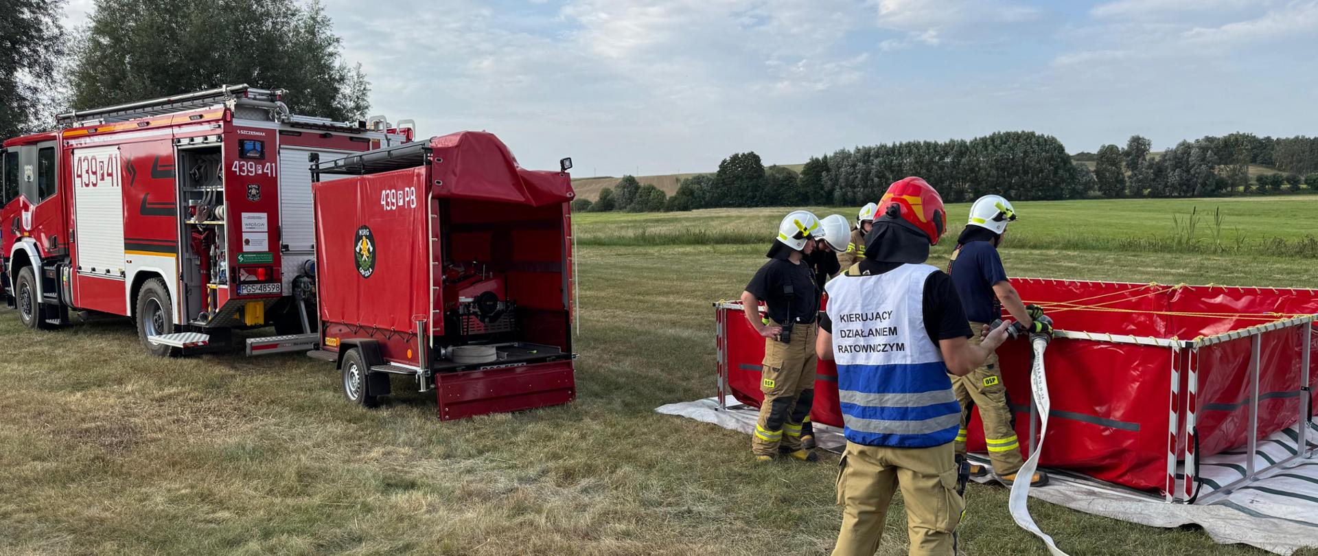 Druhów stojących przy zbiorniku wodnym. Czekają oni na podanie wody i napełnienie zbiornika. Przed nimi stoi strażak ubrany w kamizelke: Kierujący działaniem ratowniczym. Z lewej strony stoi wóz strażacki z przyczepką gaśniczą.