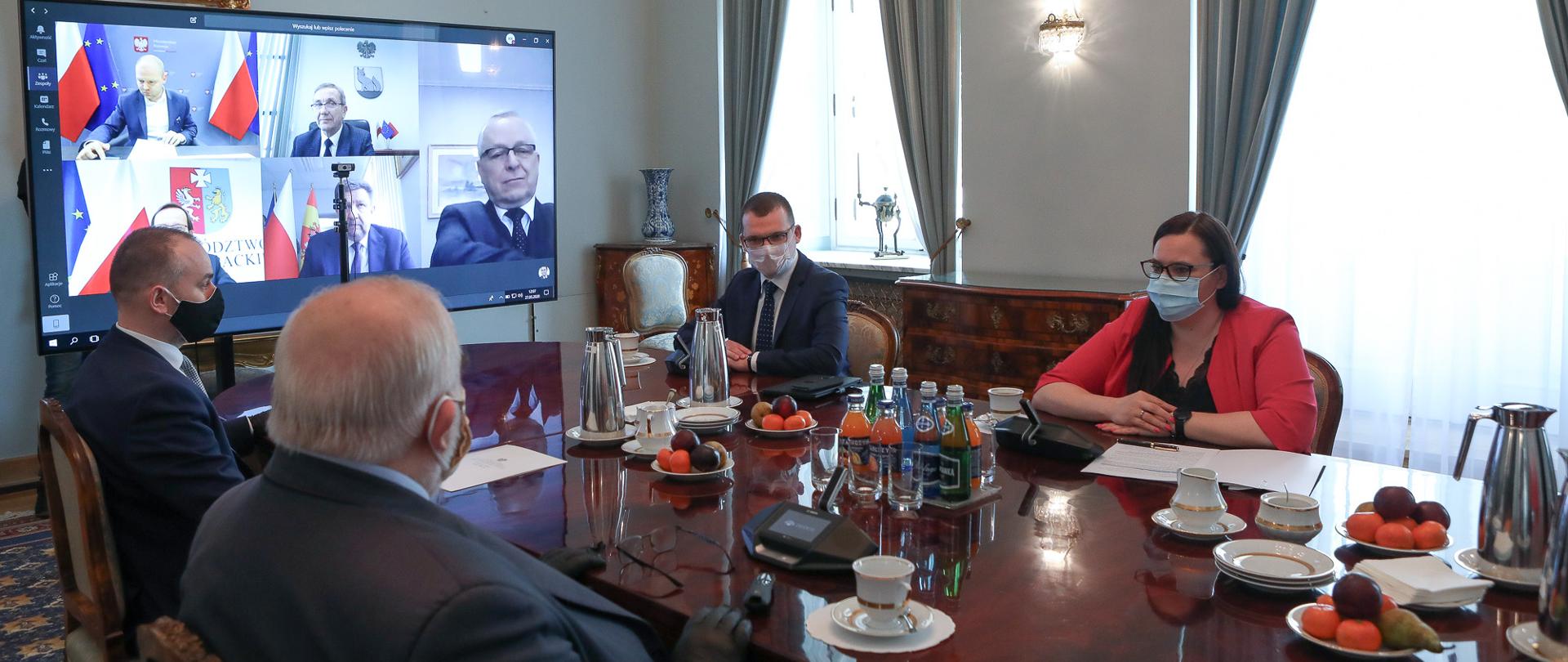 W pomieszczeniu przy dużym stole siedzą trzej mężczyźni i minister M. Jarosińska-Jedynak. Z boku na ścianie wisi monitor, na którym widać 5 osób. Trwa telekonferencja.