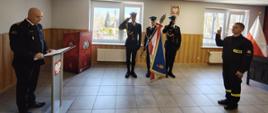 Na zdjęciu widzimy uroczystą ceremonię ślubowania strażaka. Po prawej stronie mężczyzna w mundurze strażackim unosi dwa palce prawej dłoni, składając przysięgę. Naprzeciw niego, po lewej stronie, stoi Komendant Miejski PSP w Sosnowcu, odczytując tekst ślubowania z mównicy ozdobionej godłem Polski.
W tle znajduje się poczet sztandarowy składający się z trzech strażaków w galowych mundurach, z których jeden trzyma sztandar w barwach biało-czerwonych i niebieskich. Dwaj pozostali oddają honory poprzez salutowanie. Sala jest jasna, z oknami wpuszczającymi naturalne światło, a na ścianie wisi godło Polski. Po prawej stronie widać polską flagę na stojaku.
Całość uroczystości odbywa się w oficjalnym i podniosłym tonie, wskazując na ważny moment w karierze nowo zaprzysiężonego strażaka.