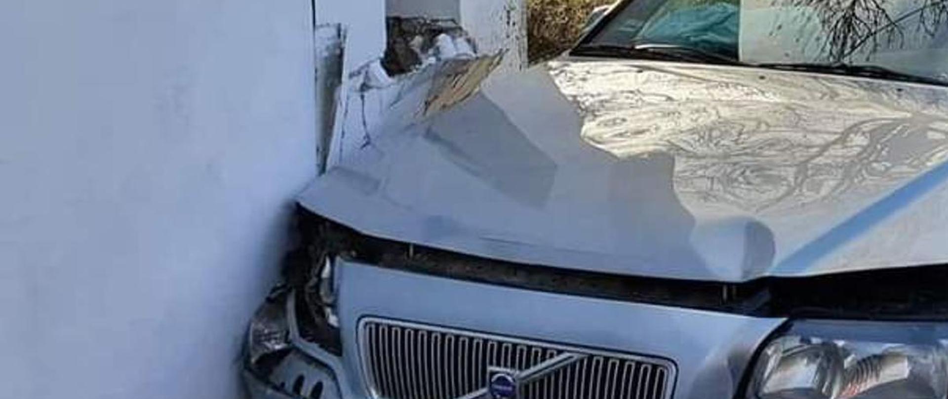 Zdjęcie przedstawia rozbite auto które uderzyło w dom oraz strażaków OSP uczestniczących w działaniach ratowniczych.