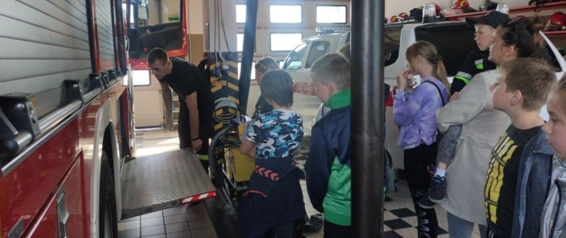 Zdjęcie wykonane wewnątrz garażu. Dzieci stoją przy wozie i słuchają strażaka ,który pokazuje sprzęt i go omawia.