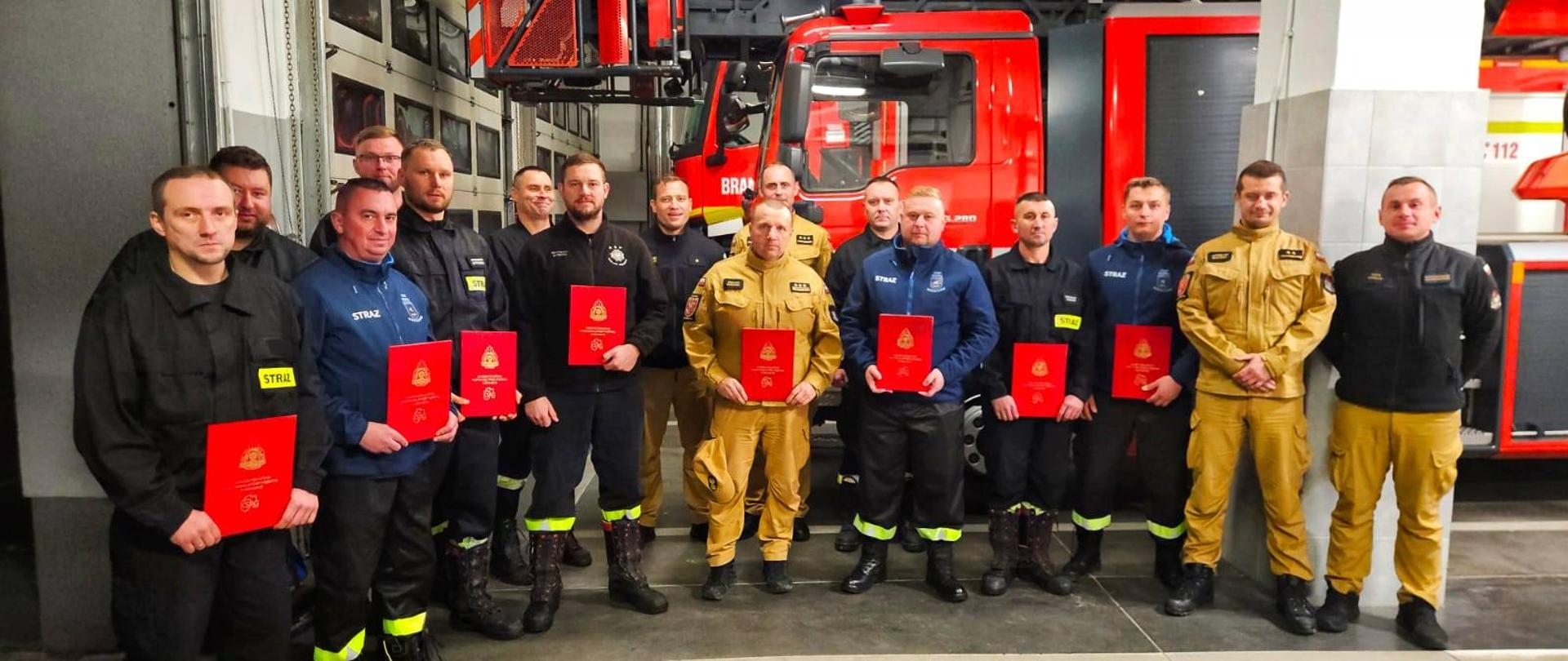 Garażu bojowym Jednostki ratowniczo Gaśniczej stoi kilkunastu strażaków. W dłoniach trzymają czerwone teczki. W tle mechaniczna drabina pożarnicza.