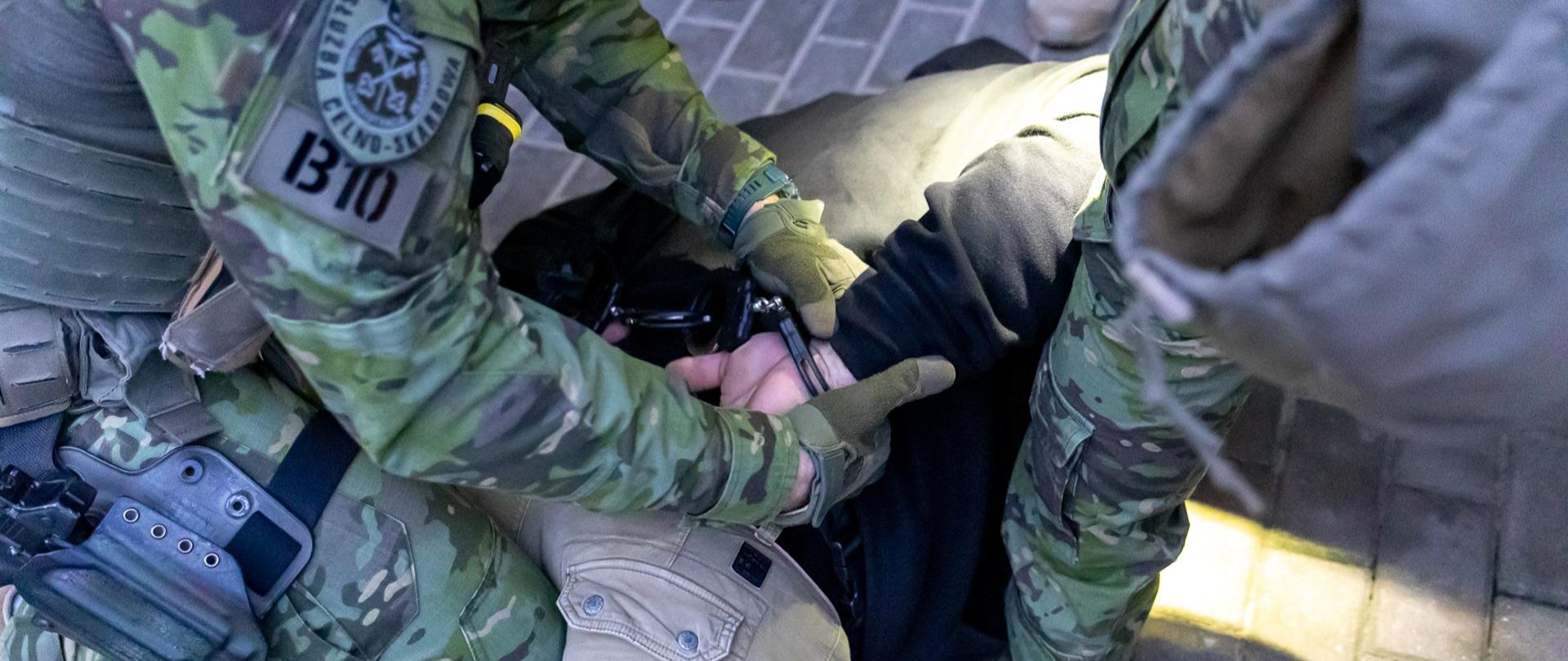 Funkcjonariusze w umundurowaniu obezwładniają osobę leżącą na chodniku i zakładają kajdanki