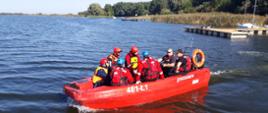 Szkolenia z ratownictwa na obszarach wodnych. Grupa strażaków ubrana w skafandry nurkowe i kamizelki ratunkowe płynie łodzią motorową po jeziorze. W tle jezioro i drzewa.