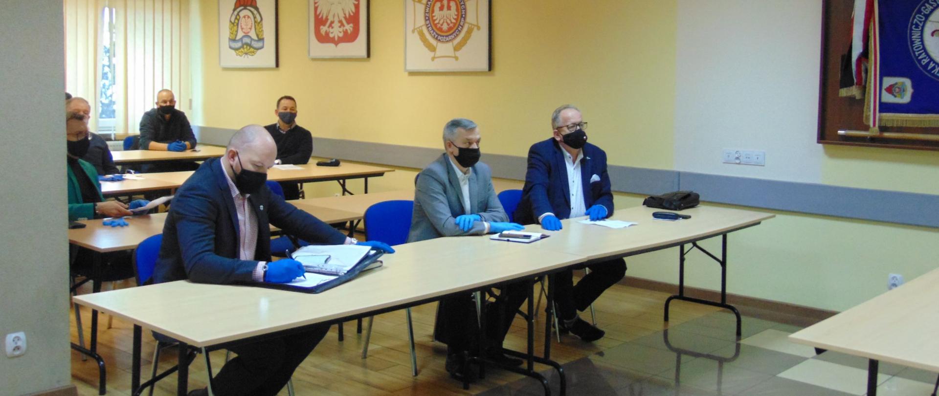 Na sali wykładowej przy ławkach siedzą mężczyźni. Wszyscy są w maseczkach i maja rękawiczki na dłoniach. Przed nimi leżą notatki.