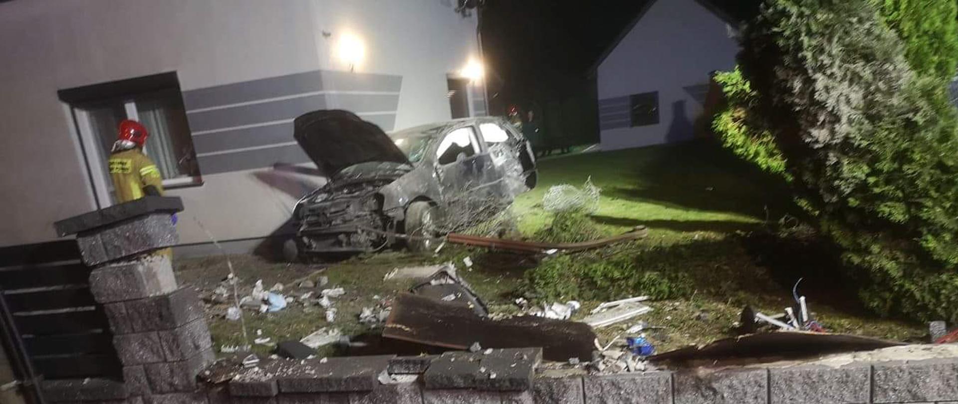 Zdjęcie przedstawia uszkodzony płot posesji oraz samochód znajdujący się przy ścianie domu. Obok stoi strażak