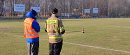 Na zdjęciu widzimy instruktora, który nadzoruje strażaka podczas zajęć praktycznych z obsługi dronu. W tle widzimy trawiaste boisko do piłki nożnej.