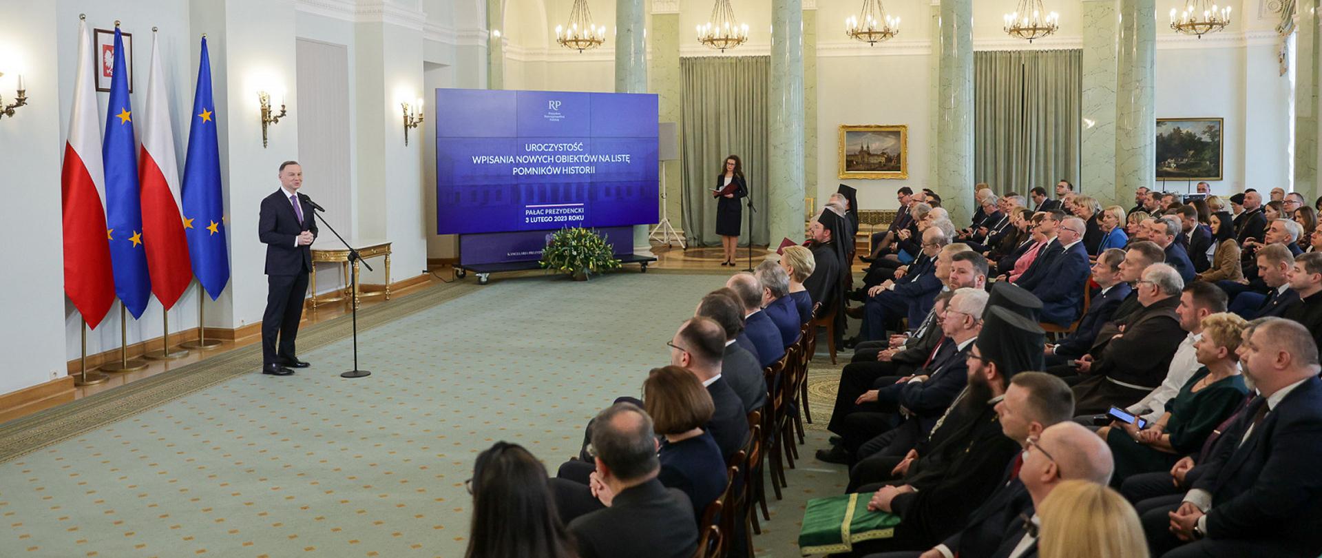 Widok z boku na dużą salę, z lewej pod flagami Polski i UE stoi prezydent Duda i mówi do mikrofonu na stojaku, z prawej na ustawionych w rzędy krzesłach siedzi dużo ludzi. W tle kolumny i żyrandole wiszące z sufitu.