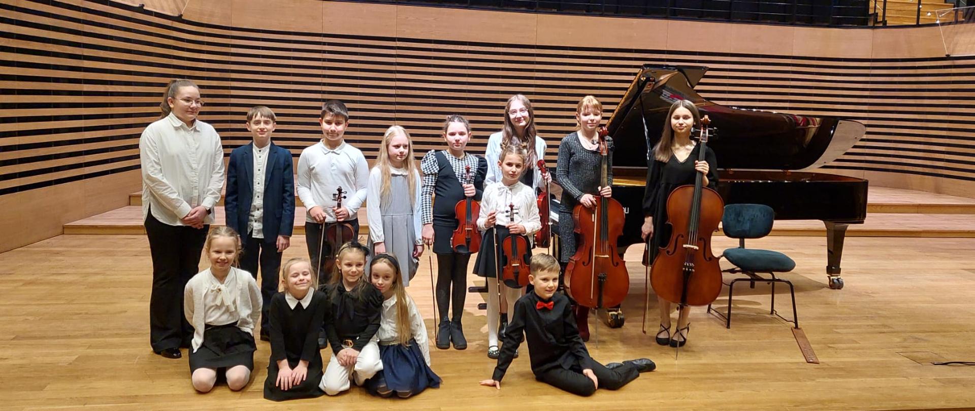 Wszyscy wykonawcy ze swoimi instrumentami stoją lub siedzą na scenie przed fortepianem.