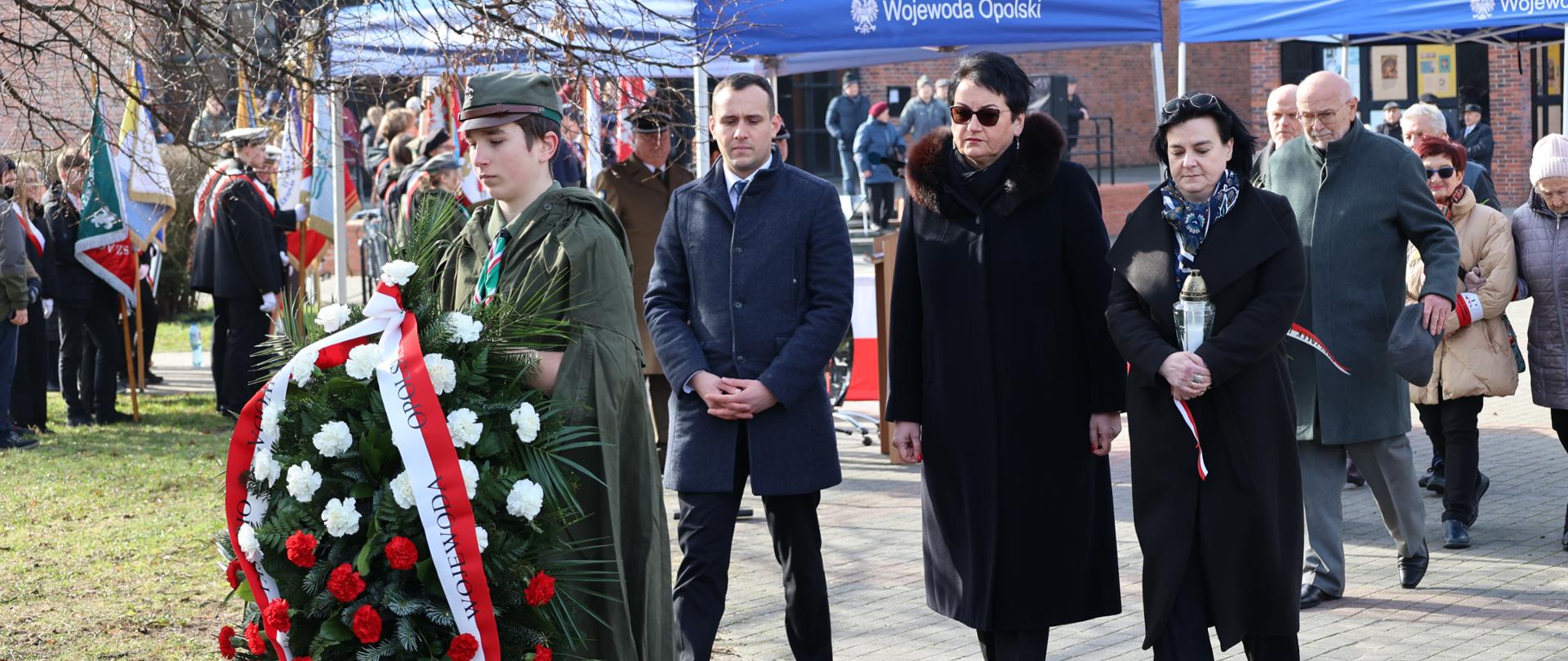 Na zdjęciu Wojewoda Opolski Monika Jurek składa kwiaty z okazji obchodów 82. Rocznicy przemianowania Związku Walki Zbrojnej w Armię Krajową. Idzie razem z Kurator Oświaty oraz Dyrektorem Biura Wojewody. Pani Kurator Oświaty niesie znicz. Przed nimi harcerz niesie składane, biało-czerwone kwiaty. 