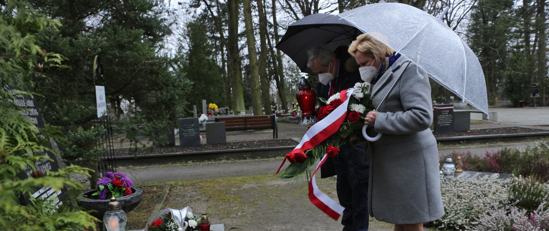 Na zdjęciu po prawej stronie wicewojewoda opolski stojąca obok wojewody opolskiego. Stoją na przeciw pomnika Golgota Wschodu w Opolu. Pod pomnikiem kwiaty i znicze - na pamiątkę wydarzeń z 1940 r. Wojewoda i wicewojewoda są ubrani w grube kurtki zimowe. Na dalszym planie groby - zdjęcie zrobiono na cmentarzu. 
