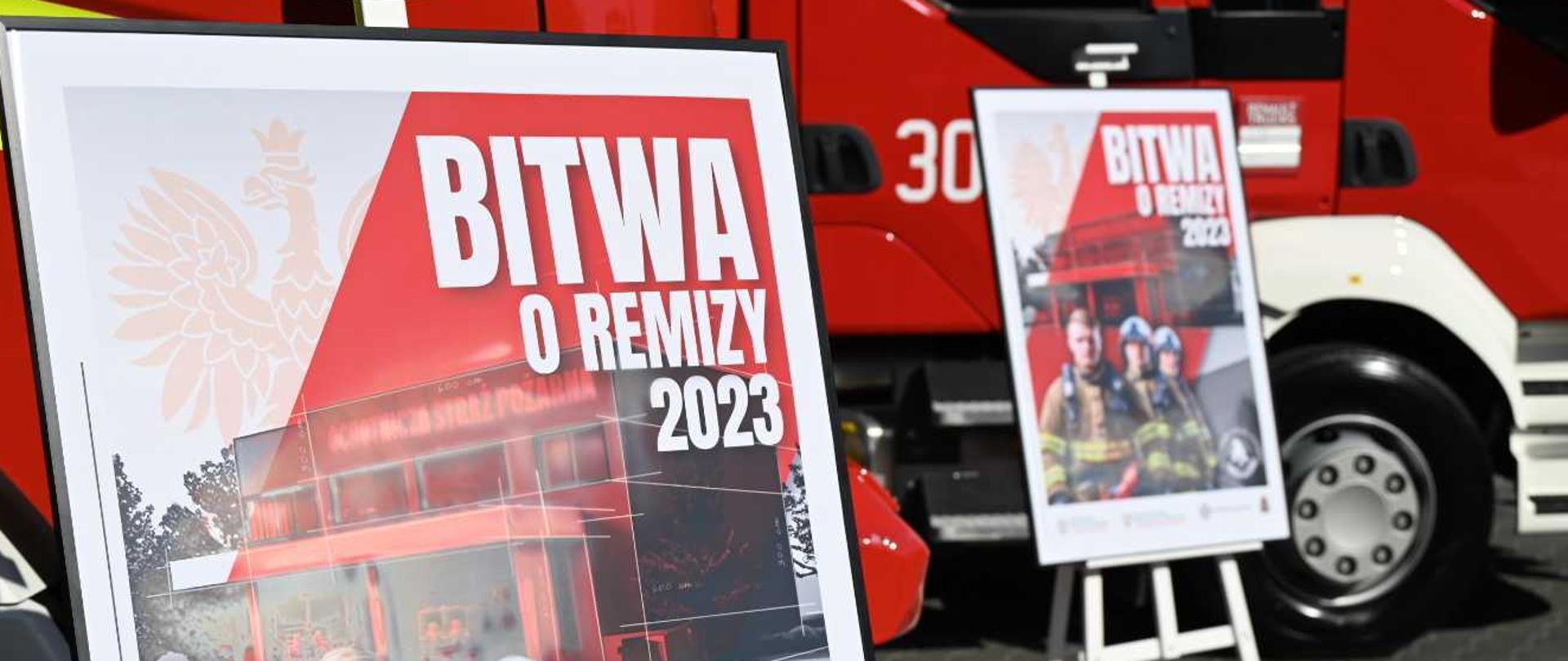 Zdjęcie przedstawia dwa plakaty promocyjne akcji "BITWA O REMIZY 2023", stojące na tle czerwonego wozu strażackiego. Lewy plakat, większy i bliżej widza, przedstawia stylizowaną grafikę remizy strażackiej w czerwono-białej kolorystyce, z orłem białym (polskim godłem) w tle. Duży, biały napis "BITWA O REMIZY 2023" dominuje na plakacie. Prawy plakat, mniejszy i umieszczony na sztaludze, również prezentuje hasło "BITWA O REMIZY 2023", ale zawiera inne elementy graficzne. W górnej części widoczna jest sylwetka remizy, a poniżej zdjęcie trzech strażaków w pełnym umundurowaniu, stojących w akcji.
Tłem dla plakatów jest fragment czerwonego wozu strażackiego z białymi elementami i numerem "30" widocznym na boku. Światło na zdjęciu wskazuje na dzień. Cała scena sugeruje wydarzenie promocyjne lub informacyjne dotyczące straży pożarnej i jej akcji.
