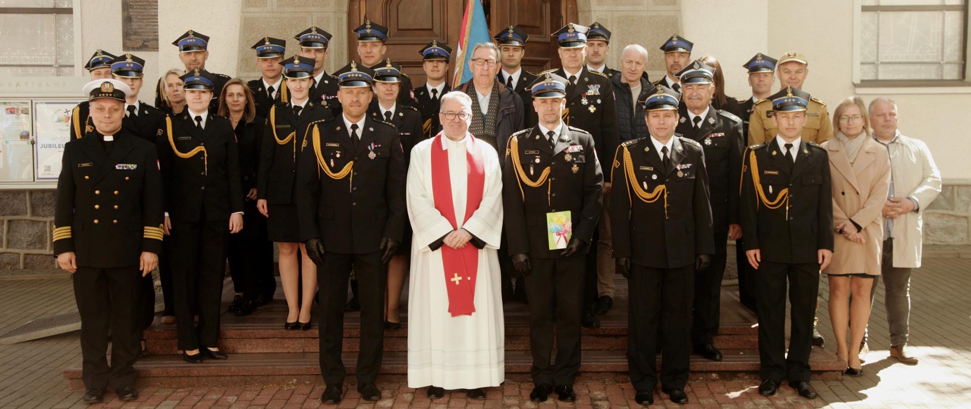 Strażacy pozujący do zdjęcia przed kościołem. Wśród nich stoi ksiądz ubrany w biało - czerwoną togę.