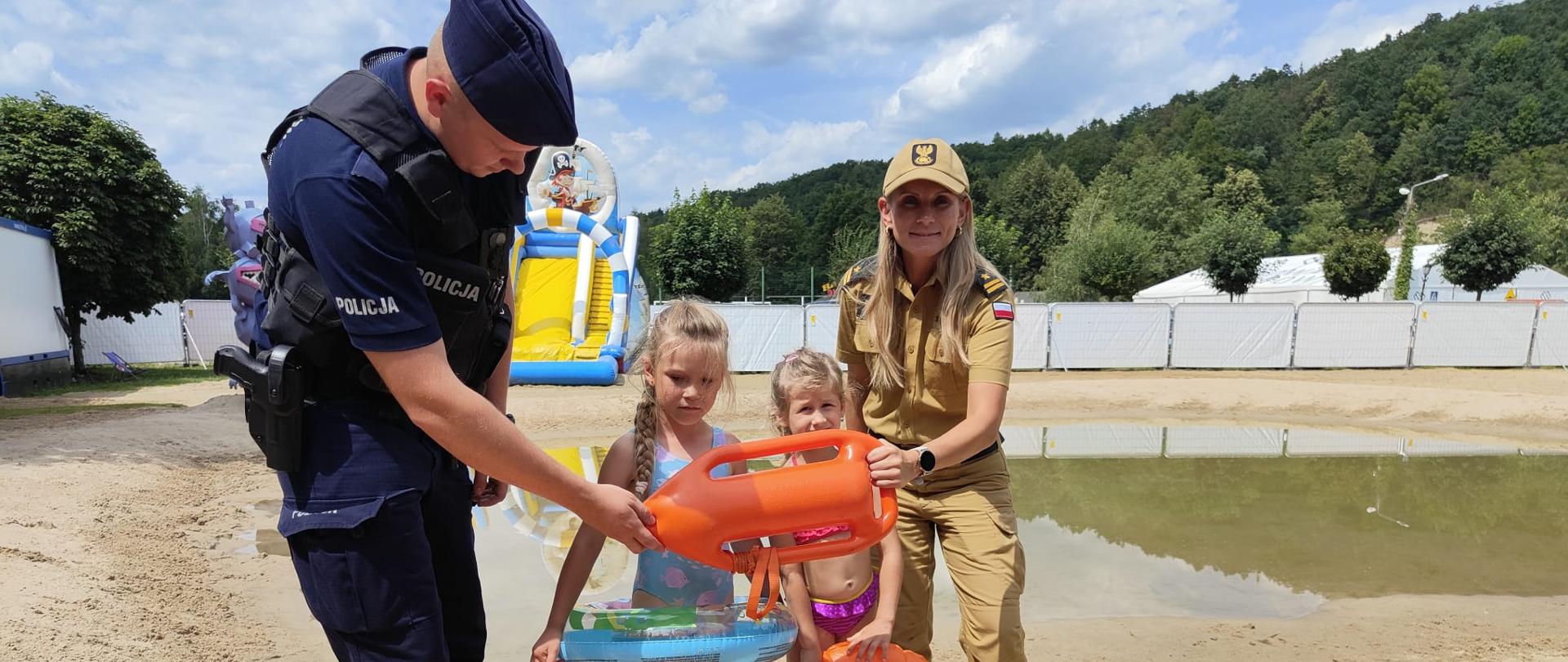 Strażak i Policjant z dwójką dzieci na plaży przy kąpielisku pokazują sprzęt do ratownictwa wodnego