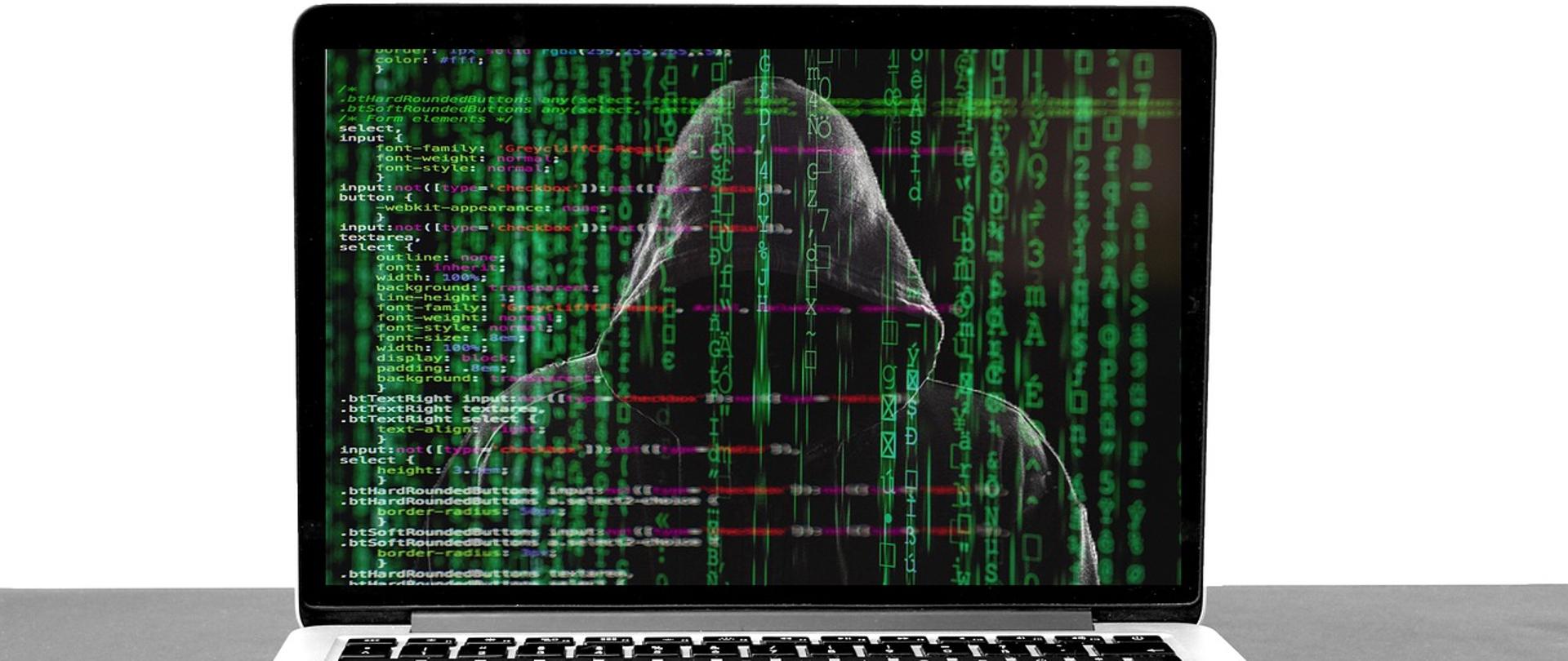 laptop, na ekranie laptopa widać postać w kapturze i dużo zielonych cyferek i literek porozrzucanych na całym ekranie
