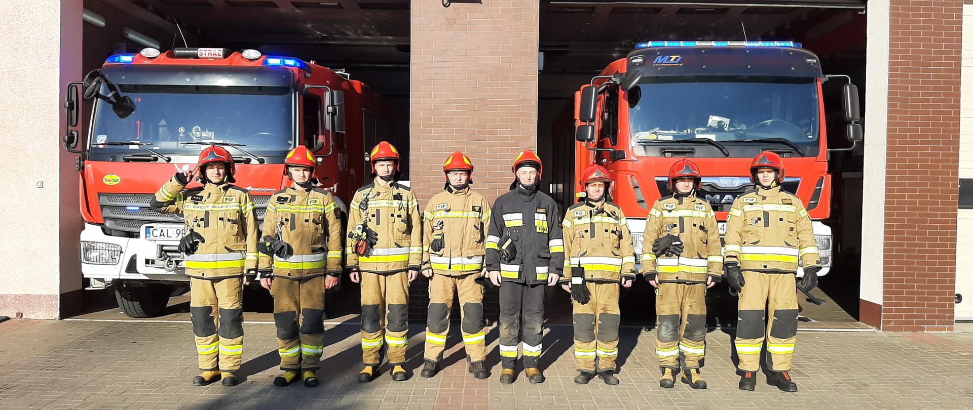 Na pierwszym planie widoczni strażacy w umundurowaniu podczas uroczystej zbiórki. W tle samochody pożarnicze i zabudowania.