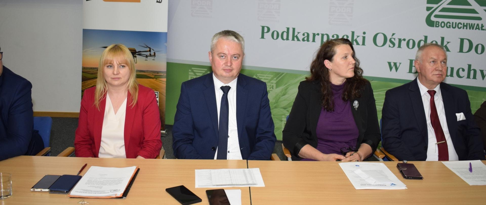 Przy długim stole konferencyjnym siedzi czworo uczestników spotkania organizowanego przez Podkarpacka Izba Rolnicza oraz Podkarpacki Ośrodek Doradztwa Rolniczego w Boguchwale. W centrum kadru znajduje się mężczyzna w granatowym garniturze i ciemnym krawacie, patrzący w stronę obiektywu. Po jego lewej stronie siedzi kobieta w czerwonej marynarce i jasnej bluzce, a po prawej kobieta w czarnym żakiecie i fioletowej bluzce. Skrajnie po prawej widoczny jest starszy mężczyzna w garniturze z bordowym krawatem. Na stole leżą dokumenty, telefony komórkowe i notatki. W tle znajdują się banery z logotypami organizatorów oraz zielono-białe tło z napisem „Podkarpacki Ośrodek Doradztwa Rolniczego w Boguchwale”.
