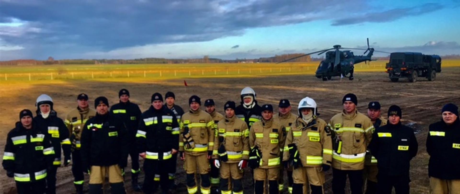 Na łące na tle śmigłowca stoją w dwuszeregu strażacy w żółtych i czarnych ubraniach bojowych.