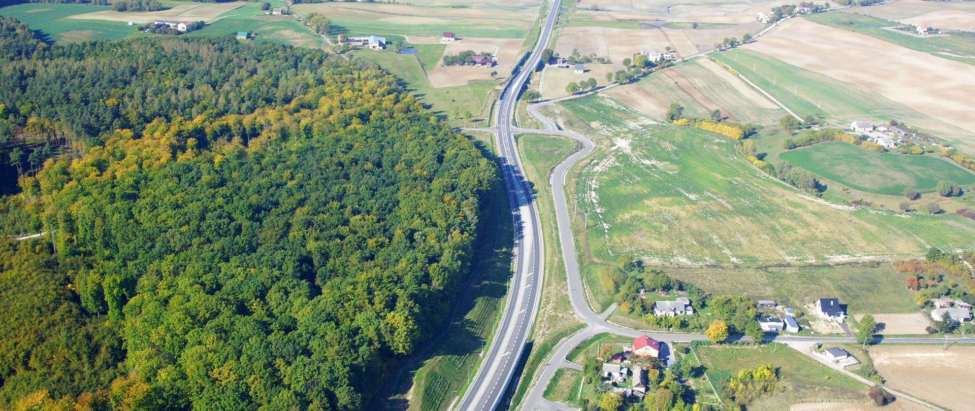 Zdjęcie przedstawia widok z lotu ptaka na drogę krajową obok drogi po lewej stronie las po prawej krajobraz rolniczy z zabudowaniami miejscowości.