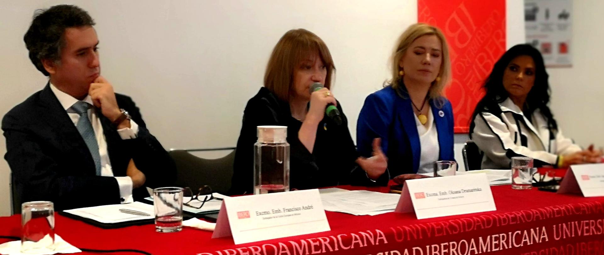 Cuatro personas sentadas a una mesa: el Embajador de la Unión Europea en México, el Embajador de Ucrania, el Embajador de Polonia y una periodista. Una de las personas habla por el micrófono. Al fondo se ve el letrero IBERO.